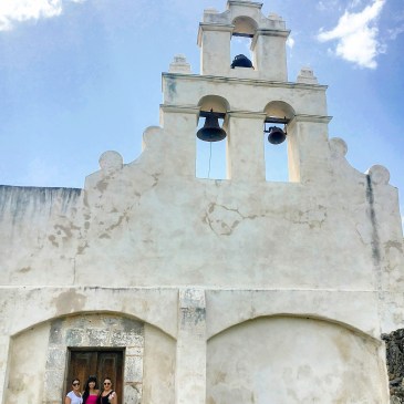 White Spanish church with three hanging bells and three Latin women standing by the door.